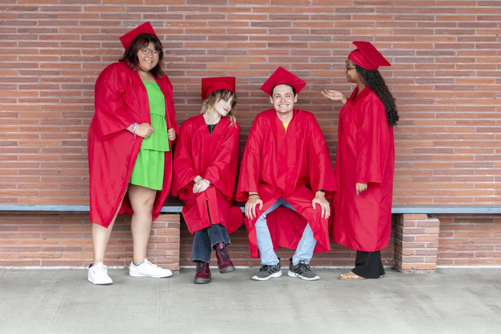 Four graduates in red caps and gowns posing casually in front of a brick wall, laughing and chatting together.
