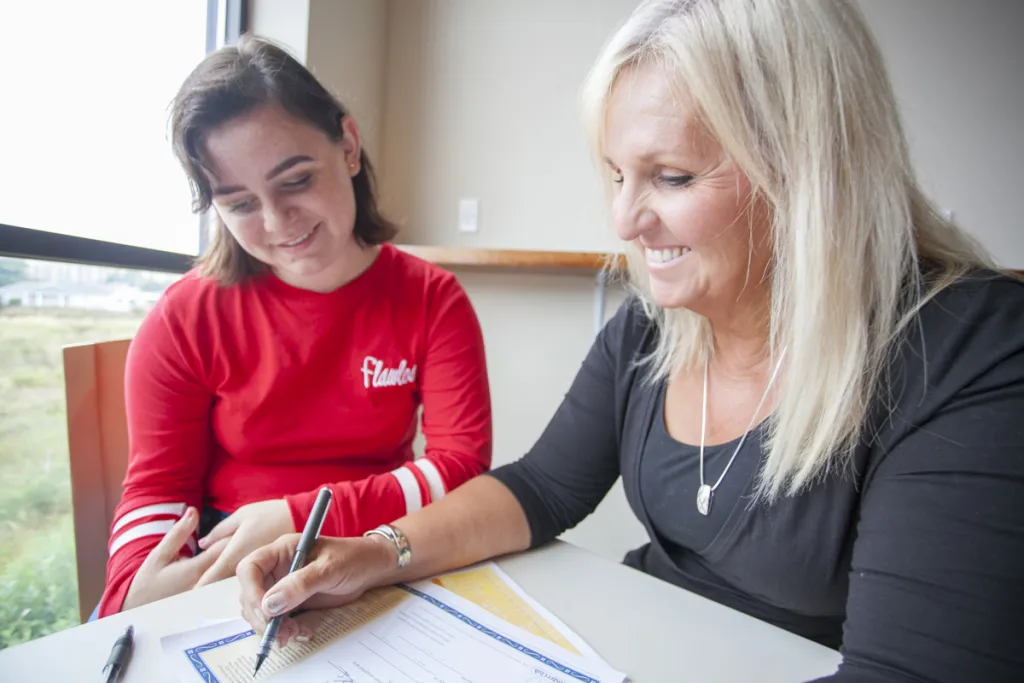 Two women smiling while reviewing and signing documents together at a table near a window.